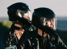 Two people wearing black helmets and leather jackets in a scenic outdoor setting.