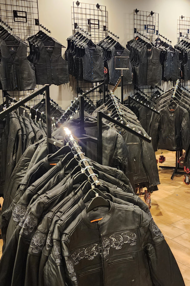 a display of black leather jackets, some with skull designs on the shoulders, arranged on racks in a store. The jackets are neatly hung, with several more displayed on the wall behind them. The lighting highlights the details of the leather and the embroidered designs.
